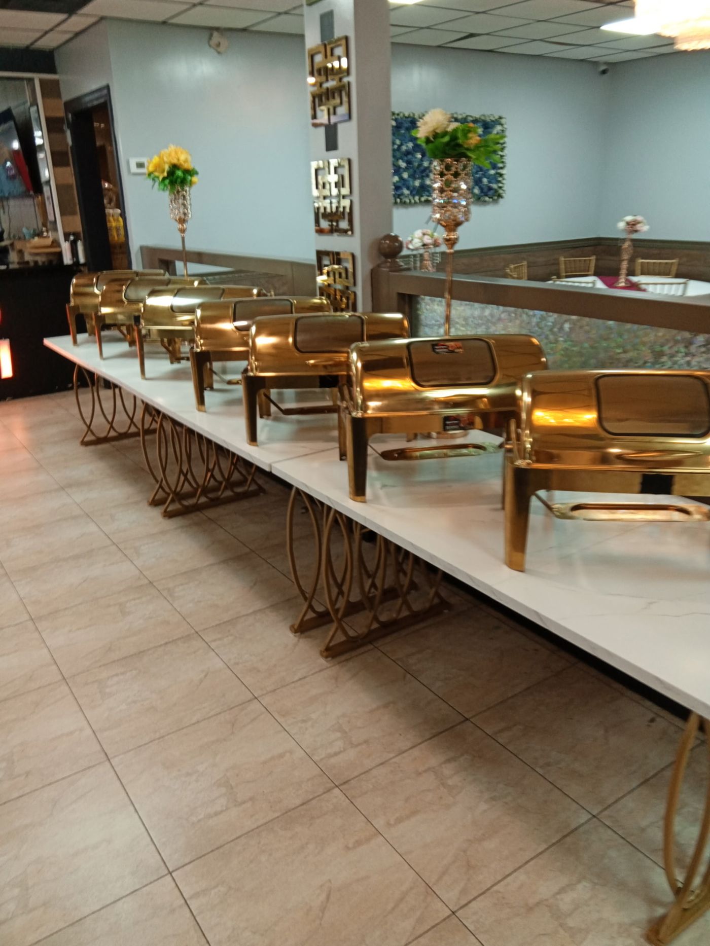 Long buffet line of polished gold chafing dishes set on white marble-top tables in a banquet hall